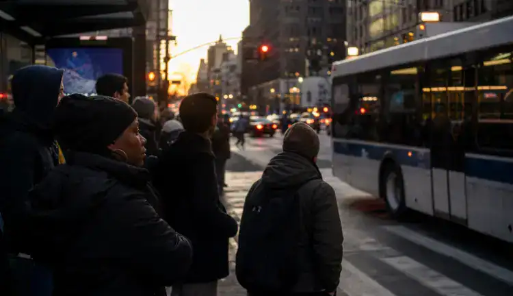 New York sidewalk at dusk near a bus stop, pedestrians waiting to cross as a bus pulls away and traffic passes.