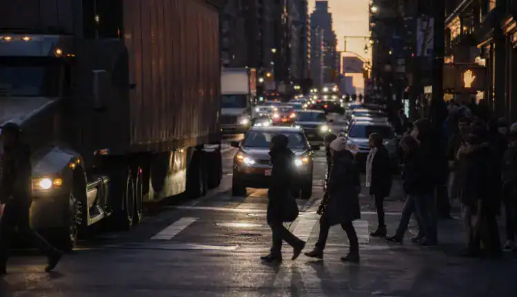 New York avenue at dusk with tractor-trailers, cars, and pedestrians waiting at a crosswalk by storefronts.