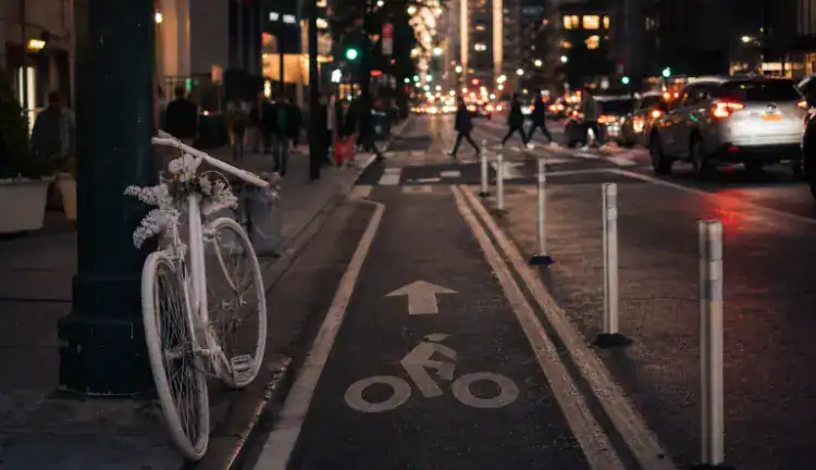 A protected bike lane on a busy New York City avenue at dusk with a ghost bike near the curb, separated by flexible bollards from slow-moving traffic and pedestrians crossing calmly in the background.