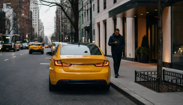 A yellow rideshare-style sedan is parked at a Manhattan curb beside a modern apartment building while a pedestrian checks a smartphone on the sidewalk and morning traffic moves past under muted city light.