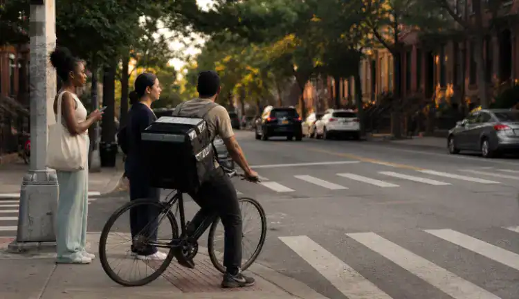 Delivery cyclist waits at a Brooklyn intersection beside pedestrians and parked cars on a typical late-afternoon street.