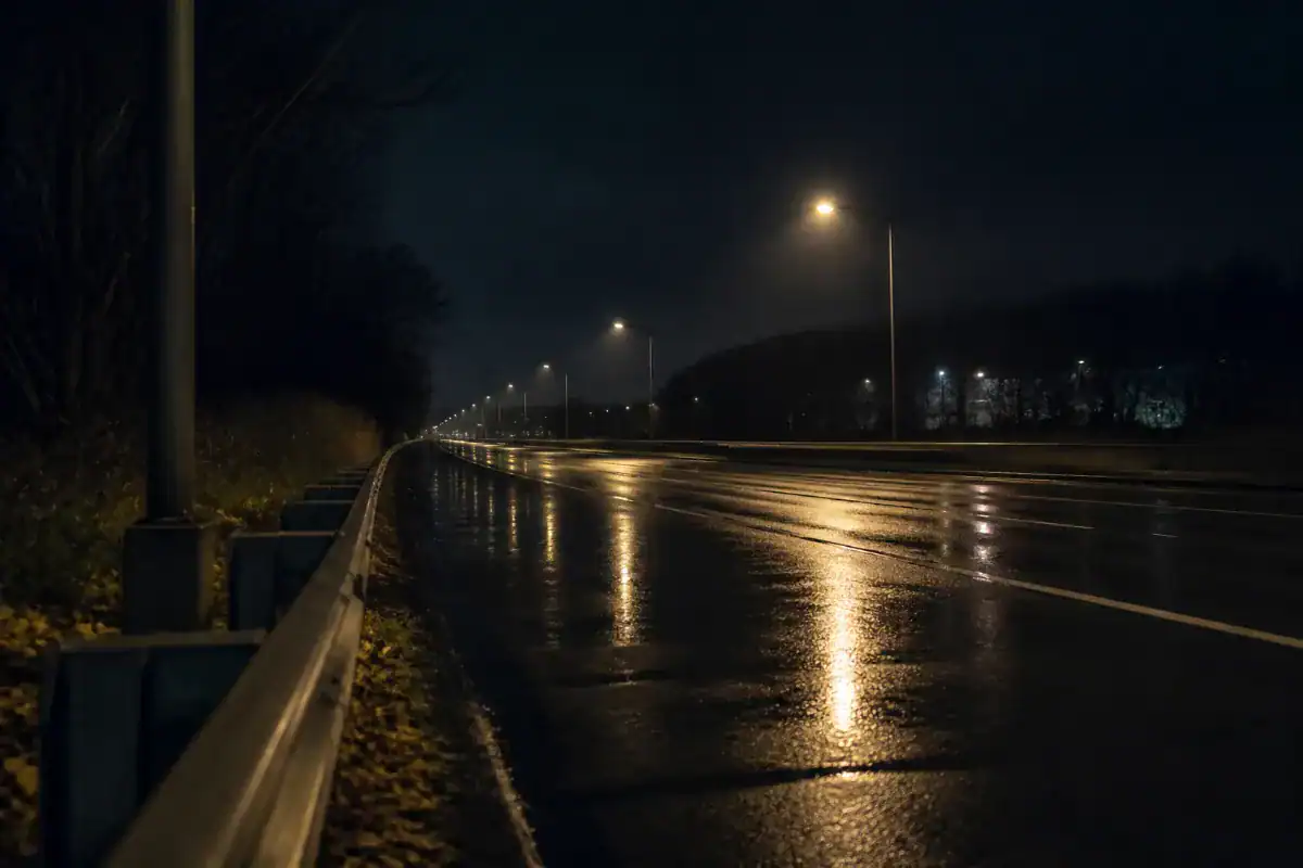 A dark rain slicked outer borough roadway at night seen from the shoulder, guardrail, streetlight reflections stretching across the wet surface, no traffic