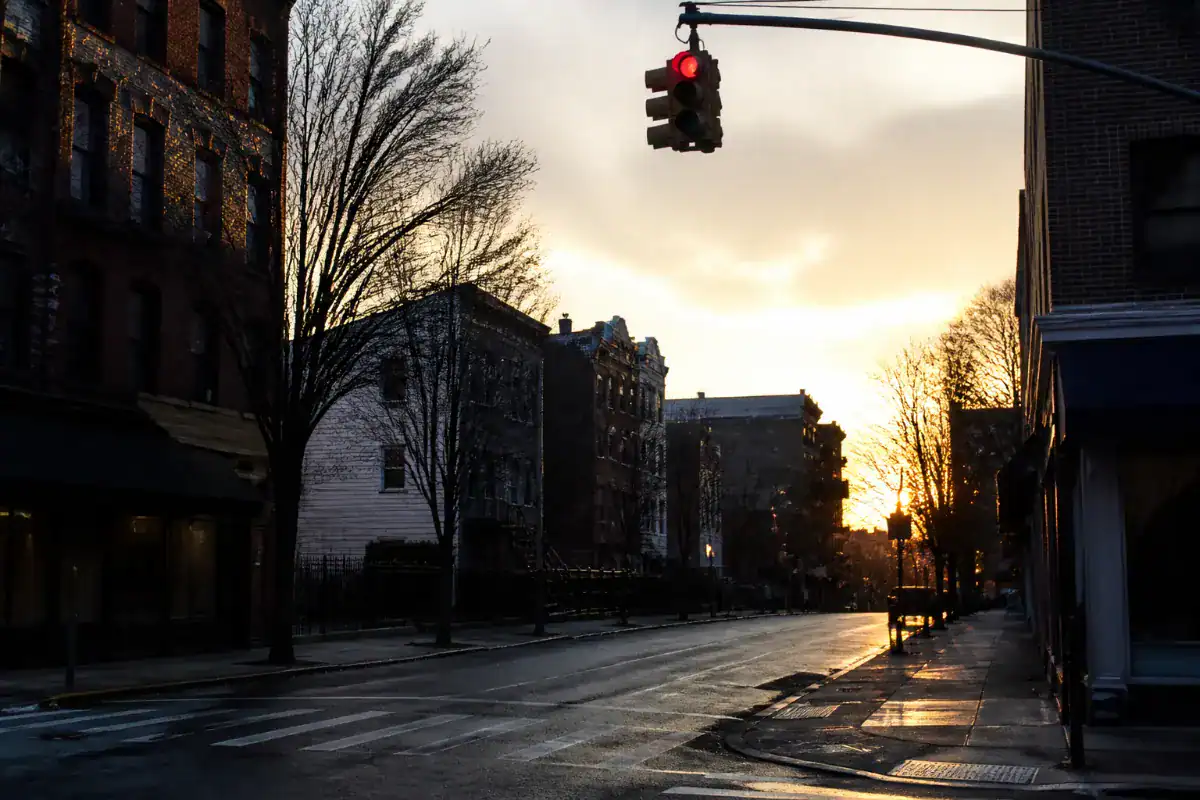 A Brooklyn intersection at dusk seen from the kerb, a traffic signal showing red, empty roadway, low winter sun behind the buildings