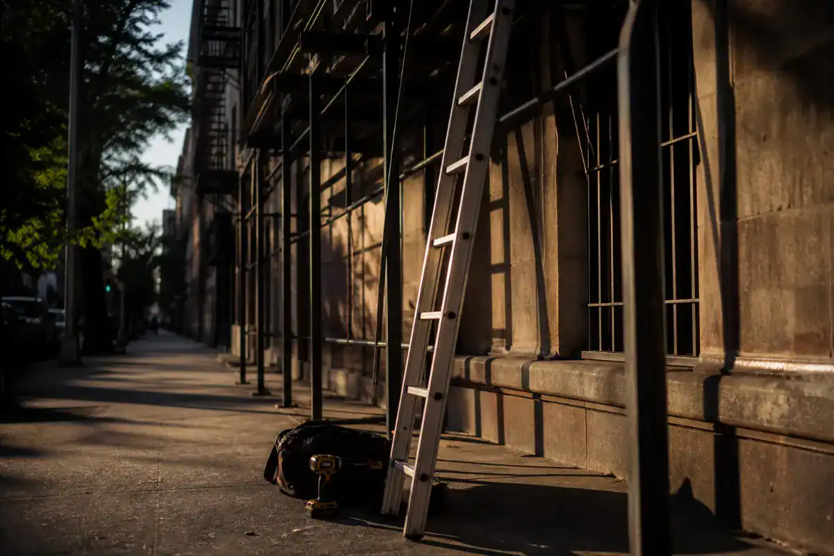 An aluminium extension ladder standing against steel scaffolding on the side of a Manhattan building, tool bag and cordless drill on the ground beside it