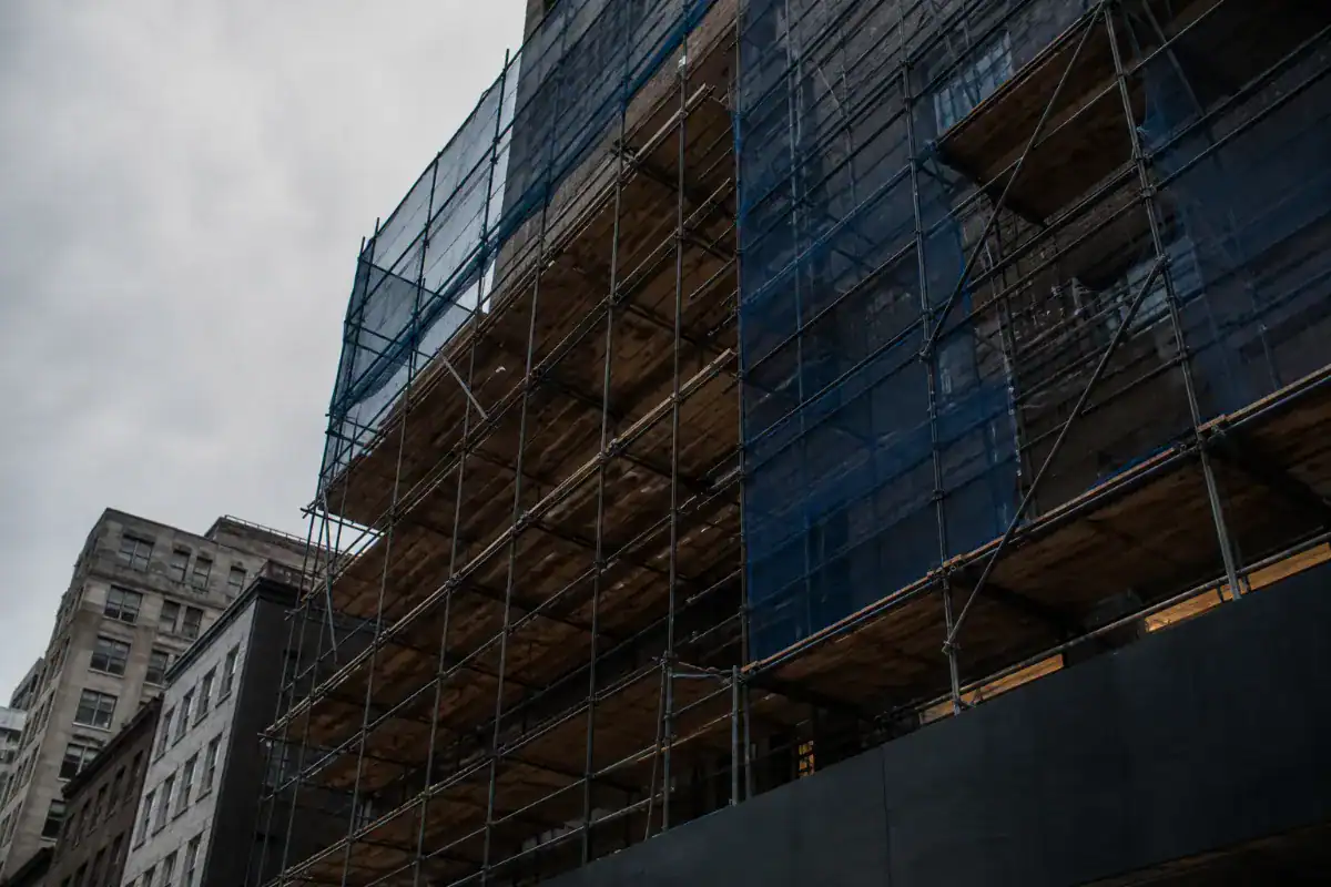 Looking up the outside of a Manhattan building at a multi level sidewalk shed and scaffolding with plywood decking and blue netting, overcast sky, no workers