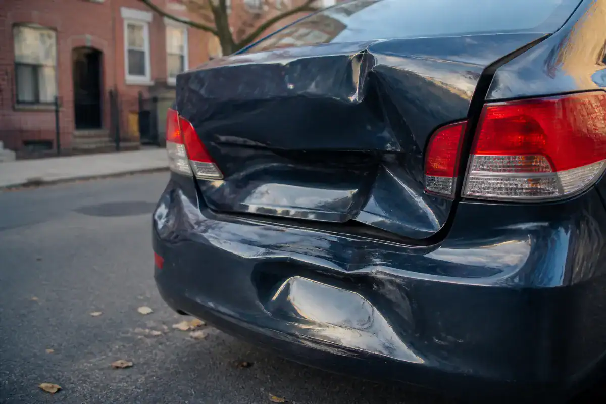 A close crop of the rear of a family saloon car with a crumpled bumper and boot lid after a rear end impact, parked on a suburban street in daylight, no people