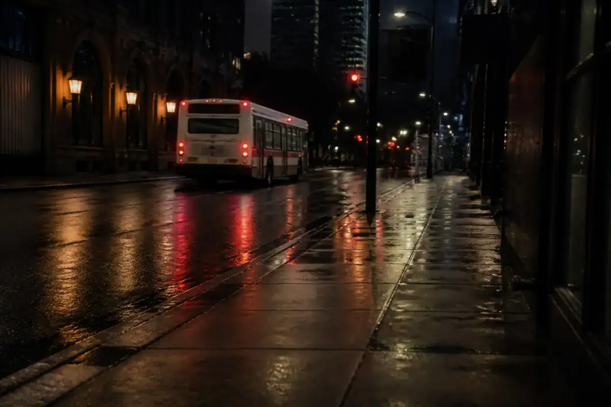 A city bus receding down a dark New York street at night with tail lights glowing, wet road, empty pavement in the foreground
