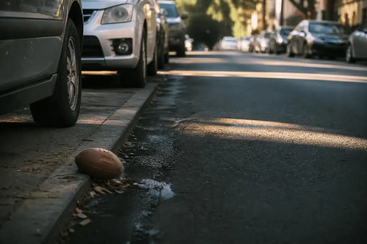 A quiet residential New York street lined with parked cars, a football resting in the gutter beside a kerb, late afternoon shadows, no people