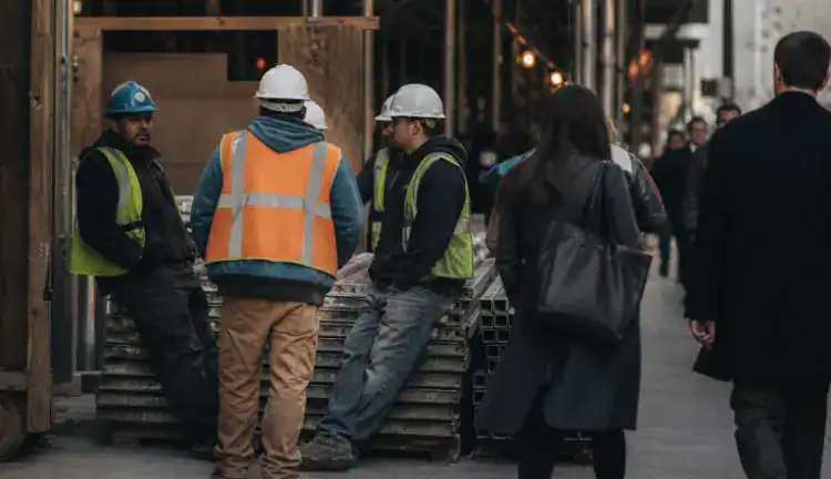 Construction workers in hard hats stand by scaffolding and stacked materials at a Manhattan site as pedestrians pass.