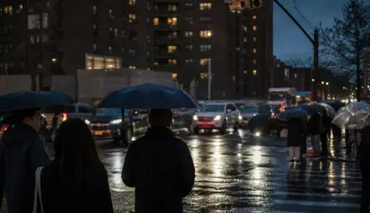 Rainy dusk at a busy Queens intersection, cars at a red light and pedestrians with umbrellas on wet pavement.