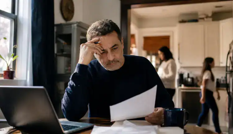 Middle-aged New Yorker at a kitchen table with papers and laptop, family in the background of a modest apartment.