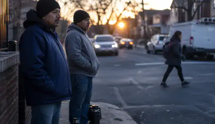 At dawn in a Queens neighborhood, two immigrant day laborers wait by a curb with thermoses as commuters pass.