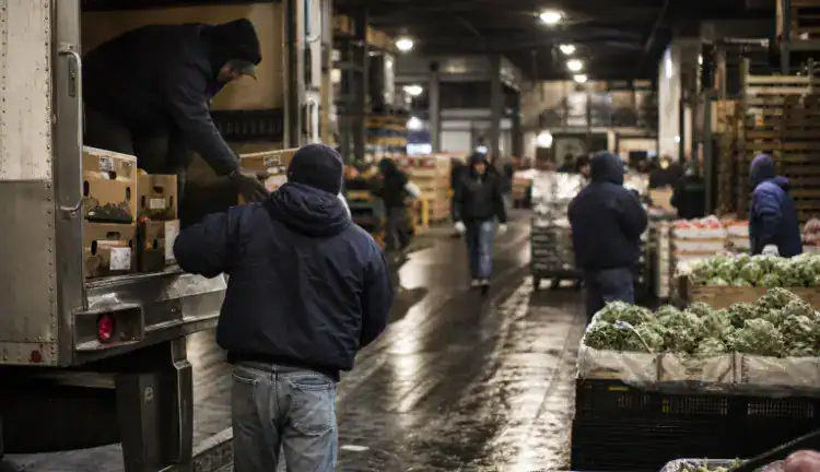 Bundled workers unload produce from a truck in a damp Queens market aisle as shoppers and vendors pass.